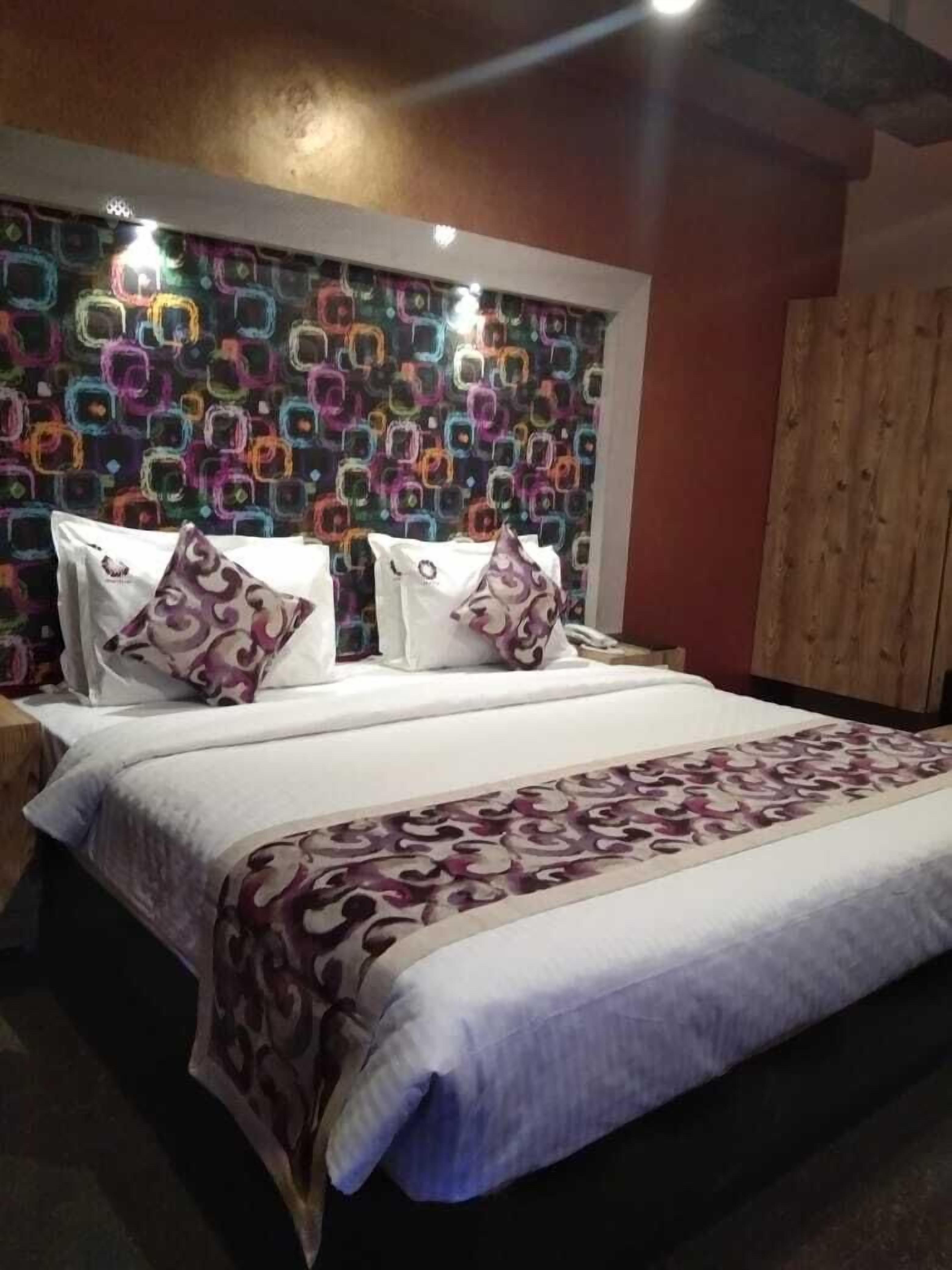 A hotel room with a patterned headboard, a large bed with a patterned bedspread, and a wooden wardrobe.