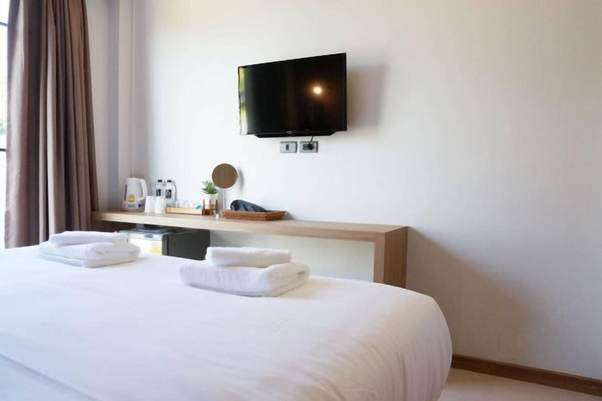 A hotel room with a large bed, a flat-screen TV mounted on the wall, a bedside table with toiletries, and a window with curtains.