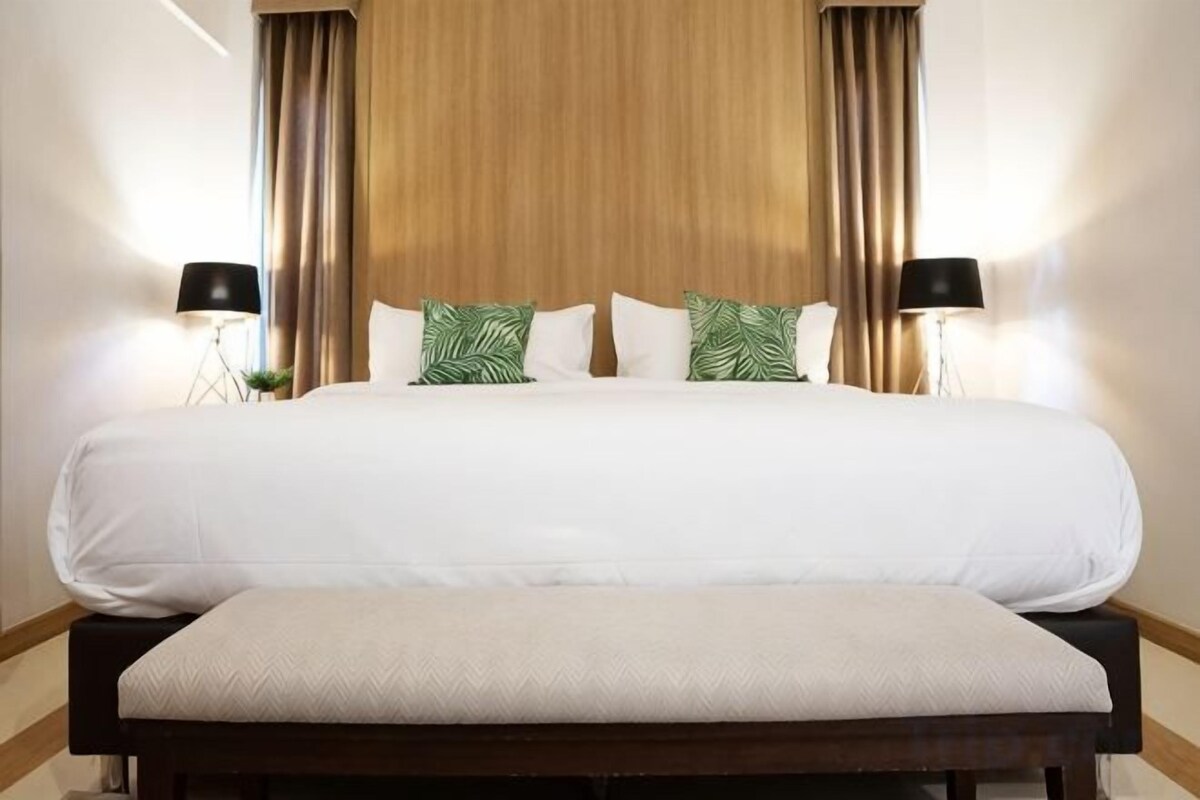 A hotel room with a large bed, a bench, two bedside lamps, and a wooden headboard.