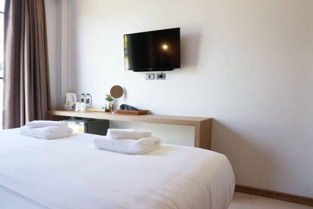 A hotel room with a bed, a flat-screen TV mounted on the wall, a bedside table with a lamp, and a window with curtains.