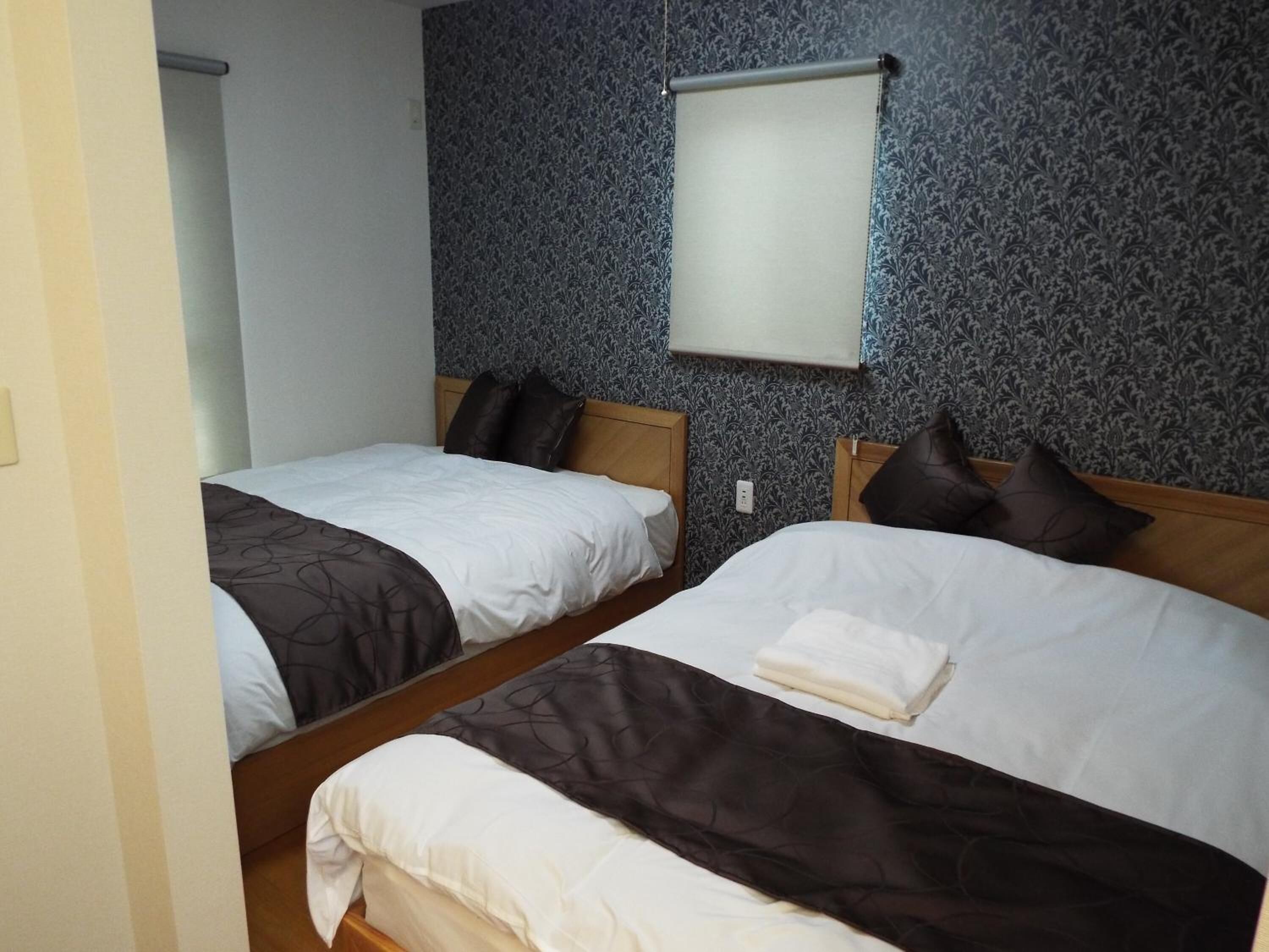 Habitación de hotel con dos camas, cada una con cabecero de madera, ropa de cama blanca y mantas marrones oscuras. Se ve una ventana pequeña con persiana en la pared.
