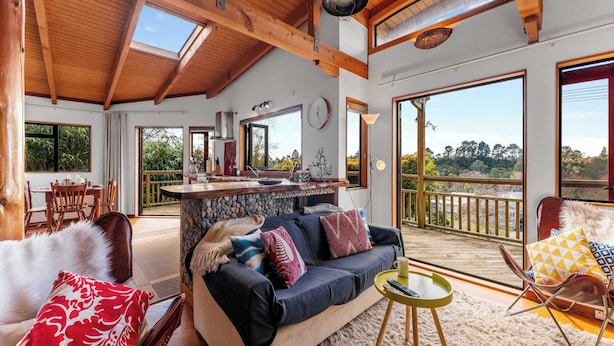 A spacious living area with a stone countertop, a sofa with colorful cushions, a dining table with chairs, and large windows offering a view of the outdoors. - Treetop Hideaway - Taupo Holiday Home (Taupo Central)