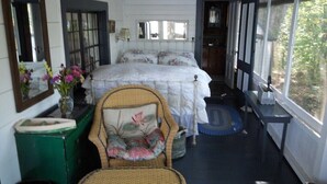 A bedroom with a large bed, a wicker chair, a green dresser, and a window with a view of trees. - Classic Lakeside Maine Cottage (Sumner)