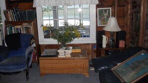 A cozy room with a wooden paneled wall, a window with a white valance, a bookshelf, a desk with books, a blue chair, and a black leather sofa. - Classic Lakeside Maine Cottage (Sumner)