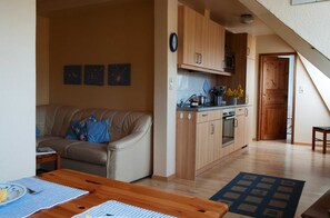 A cozy living room with a sofa and a wooden dining table, a kitchen with wooden cabinets and a microwave, and a wooden door leading to another room.