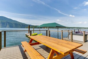 A wooden deck with picnic tables and a green umbrella, overlooking a calm lake with mountains in the background. - Waterfront house in Bayview! Boating, fishing, amazing views, 12mi to Silverwood (Bayview)