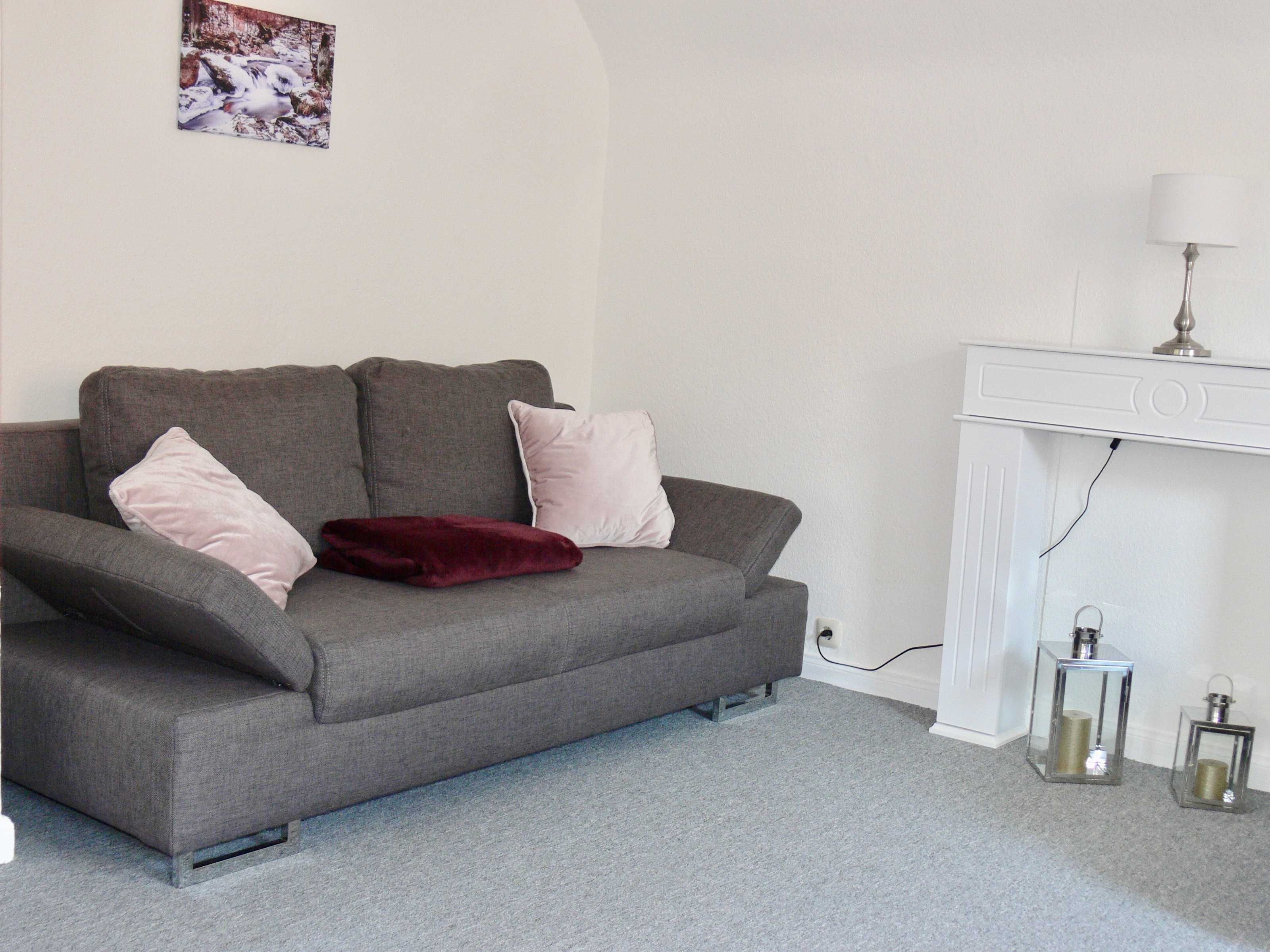 A grey sofa with two pink cushions and a red throw, a white console table with a lamp, and two glass lanterns on a grey carpet.