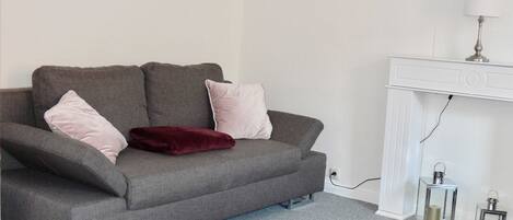 A grey sofa with two pink cushions and a red throw, a white console table with a lamp, and two glass lanterns on a grey carpet.