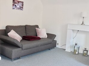 Ein graues Sofa mit zwei rosafarbenen Kissen und einer roten Decke, ein weißer Konsolentisch mit einer Lampe sowie zwei Glaskostengläser auf einem grauen Teppich.