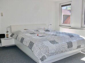Ein Schlafzimmer mit einem weißen Bett, einem Nachttisch, einer Lampe und einem Fenster mit Blick auf ein Gebäude mit rotem Dach.