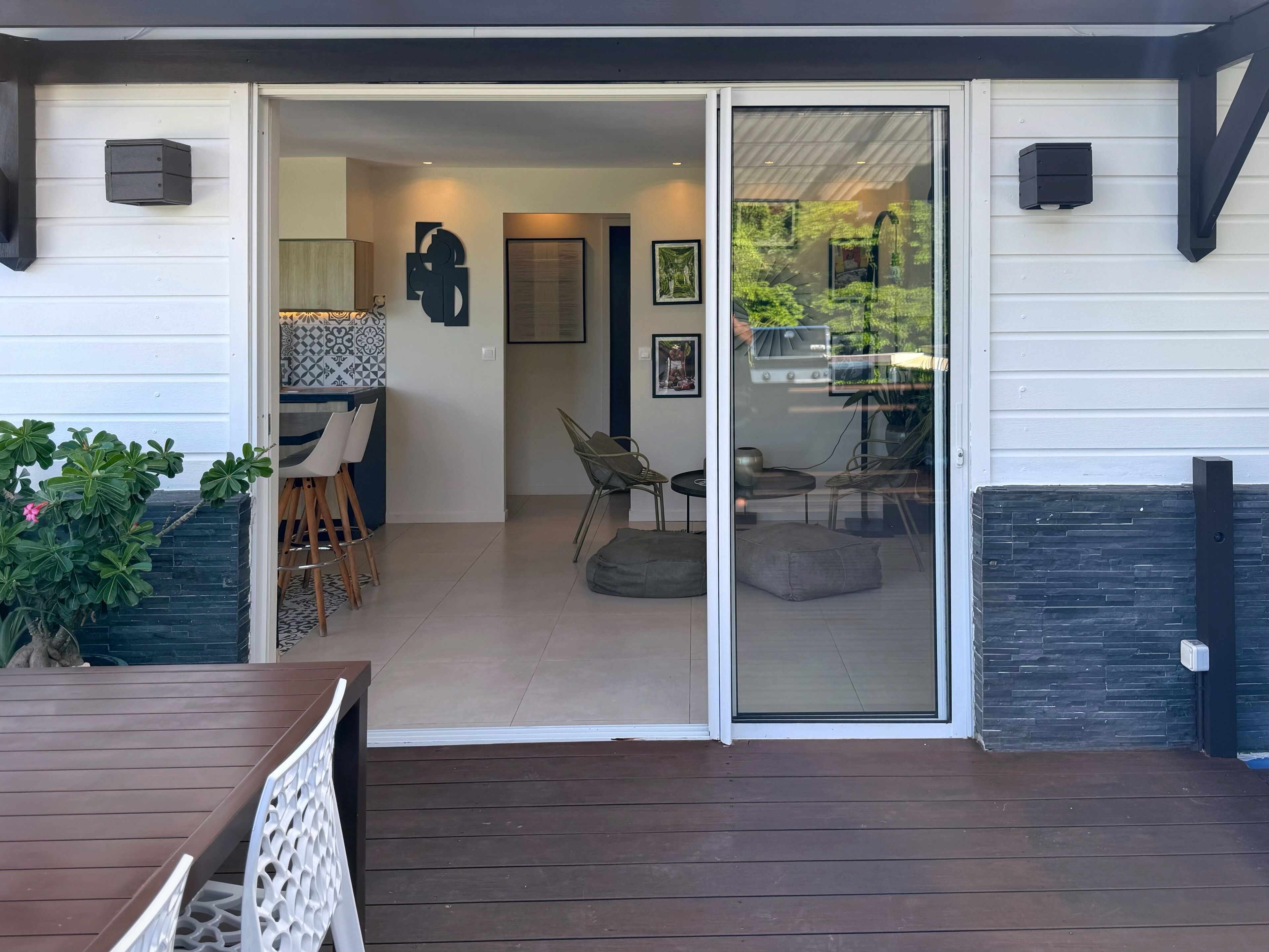 A patio with a wooden deck, a dining area with chairs and a table, and a sliding glass door leading to an indoor space with a kitchen and seating area.
