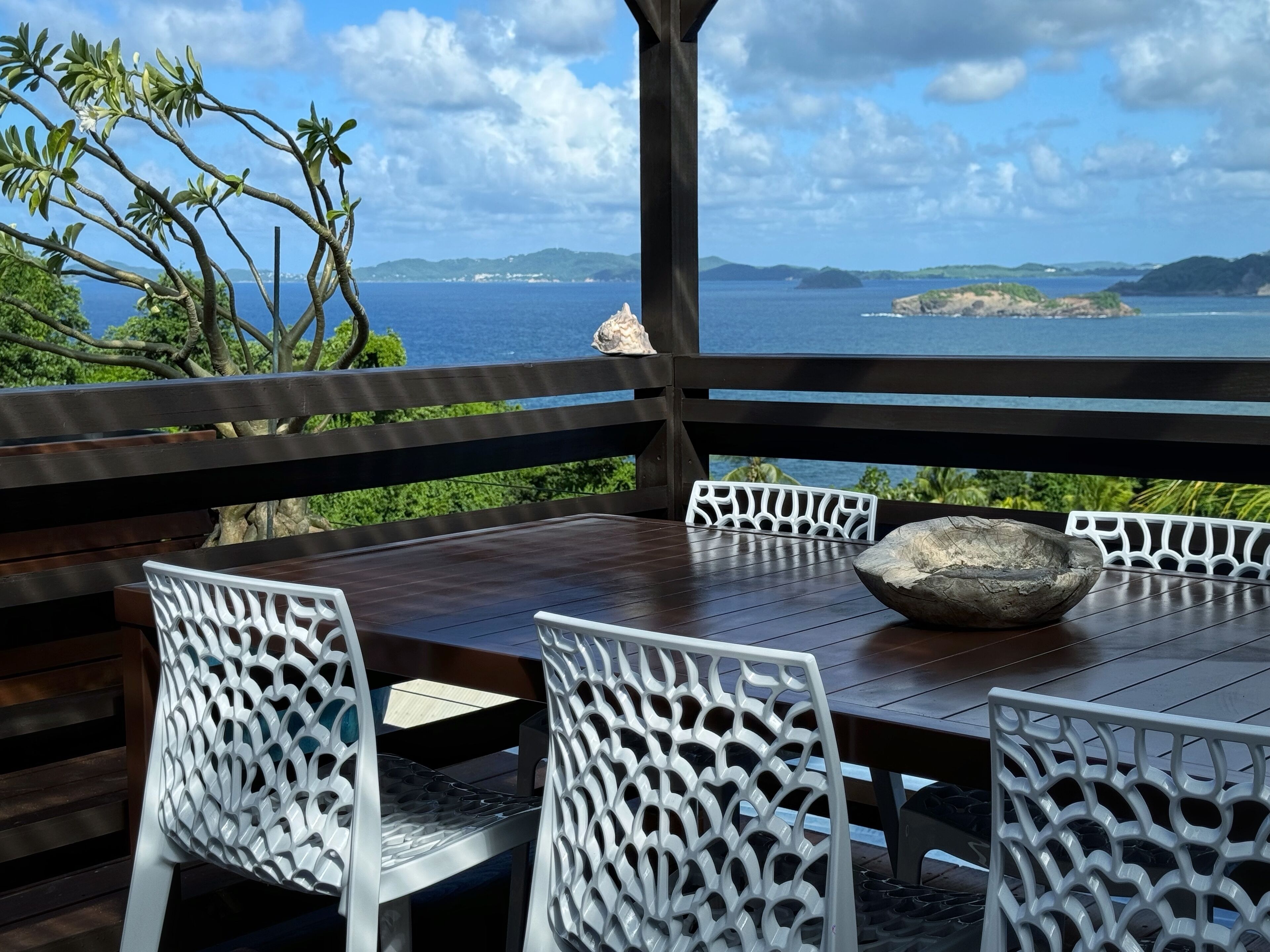 Une table en bois, entourée de chaises blanches, et un bol décoratif, le tout offrant une vue panoramique sur la mer et les îles lointaines.