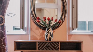 Una habitación con una mesa consola de madera, un espejo con marco decorativo, un taburete de terciopelo rojo y una ventana con cortinas.