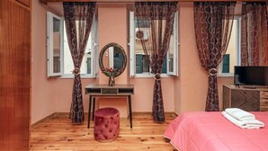 Un dormitorio con piso de madera, cama con ropa de cama rosa, un televisor sobre un mueble de madera, un otomano redondo pequeño y cortinas con estampado de hojas.