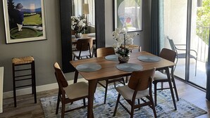 Een eetgedeelte met een houten tafel, stoelen en een kroonluchter. Er hangen afbeeldingen in lijsten aan de muur en er is een raam met gordijnen.