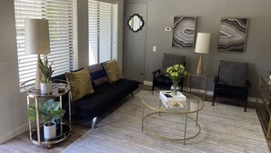 Een moderne woonkamer met een zwarte bank, een glazen salontafel en twee fauteuils. Er liggen decoratieve kussens, een vaas met bloemen en een wanddecoratie.