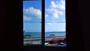 A window with a view of a coastal area, including a road, parked vehicles, and a body of water. - Hotel Henza (Uruma)