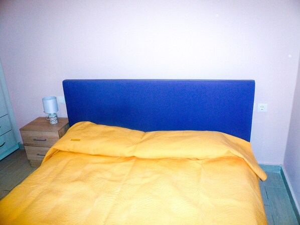 Una cama con cabecera azul, una mesita de noche de madera con una lámpara y un edredón amarillo.