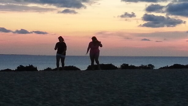 To personer står på en sandstrand med solnedgang i baggrunden.