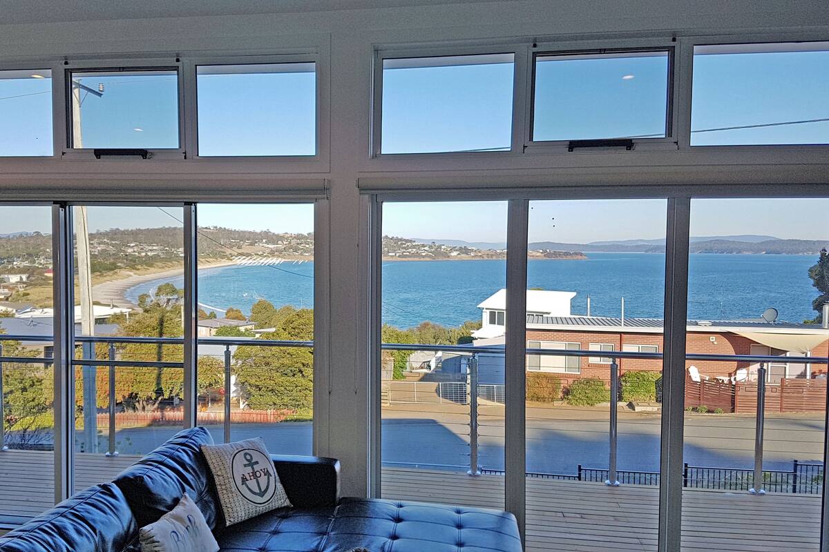 A room with a large window overlooking a coastal area, a leather sofa, and a view of the sea and distant buildings.