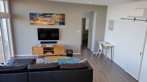 Una sala moderna con un sofá seccional grande, un televisor de pantalla plana, un mueble de madera y una zona de comedor con una mesa pequeña y sillas.