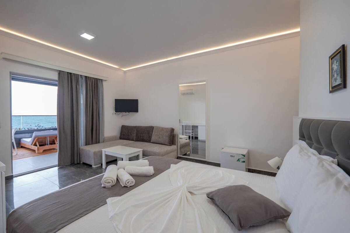 A modern hotel room with a bed, a sofa, a small table, a TV, and a view of the sea.
