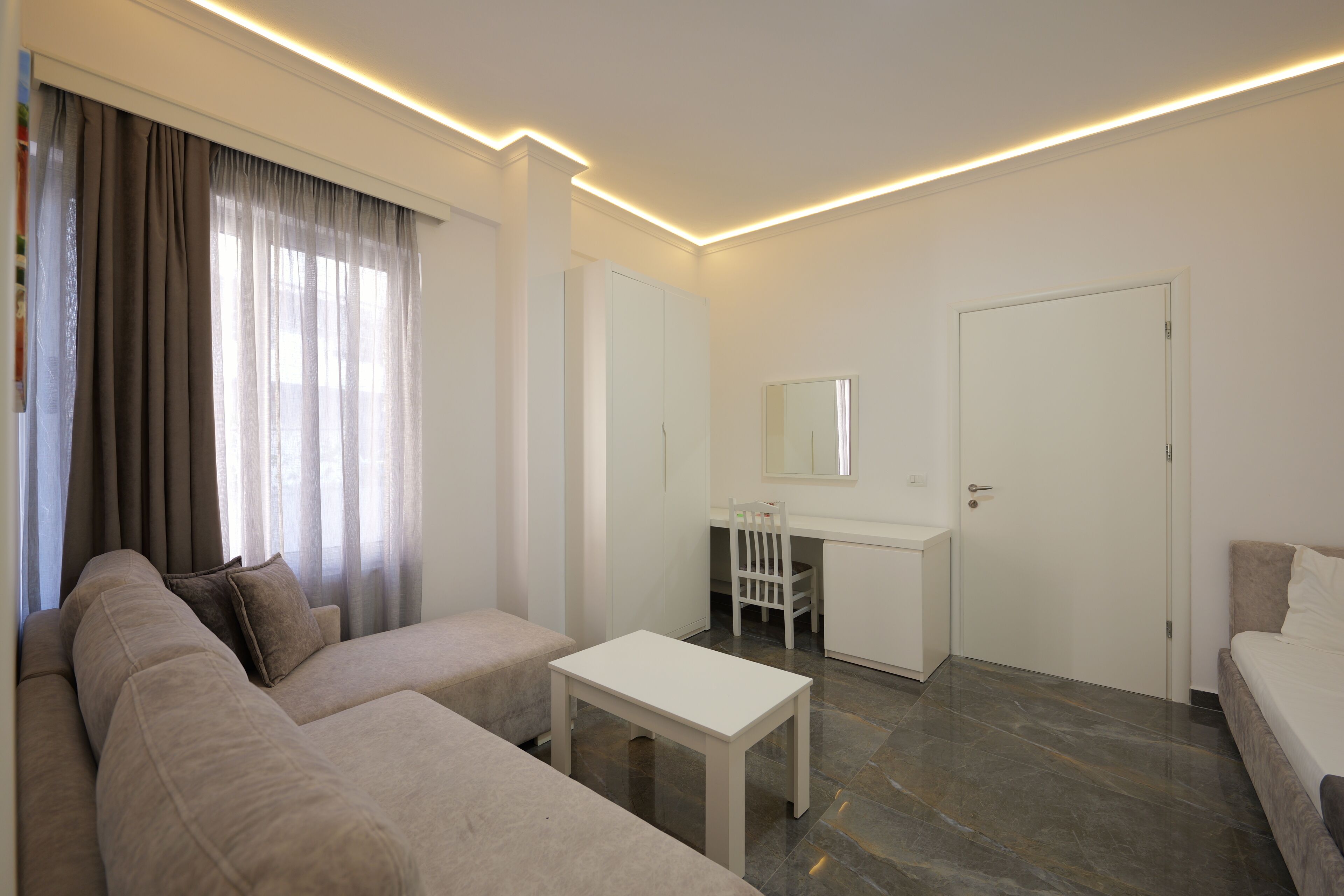 A modern hotel room with a beige sofa, a small white table, a desk with a chair, and a bed.