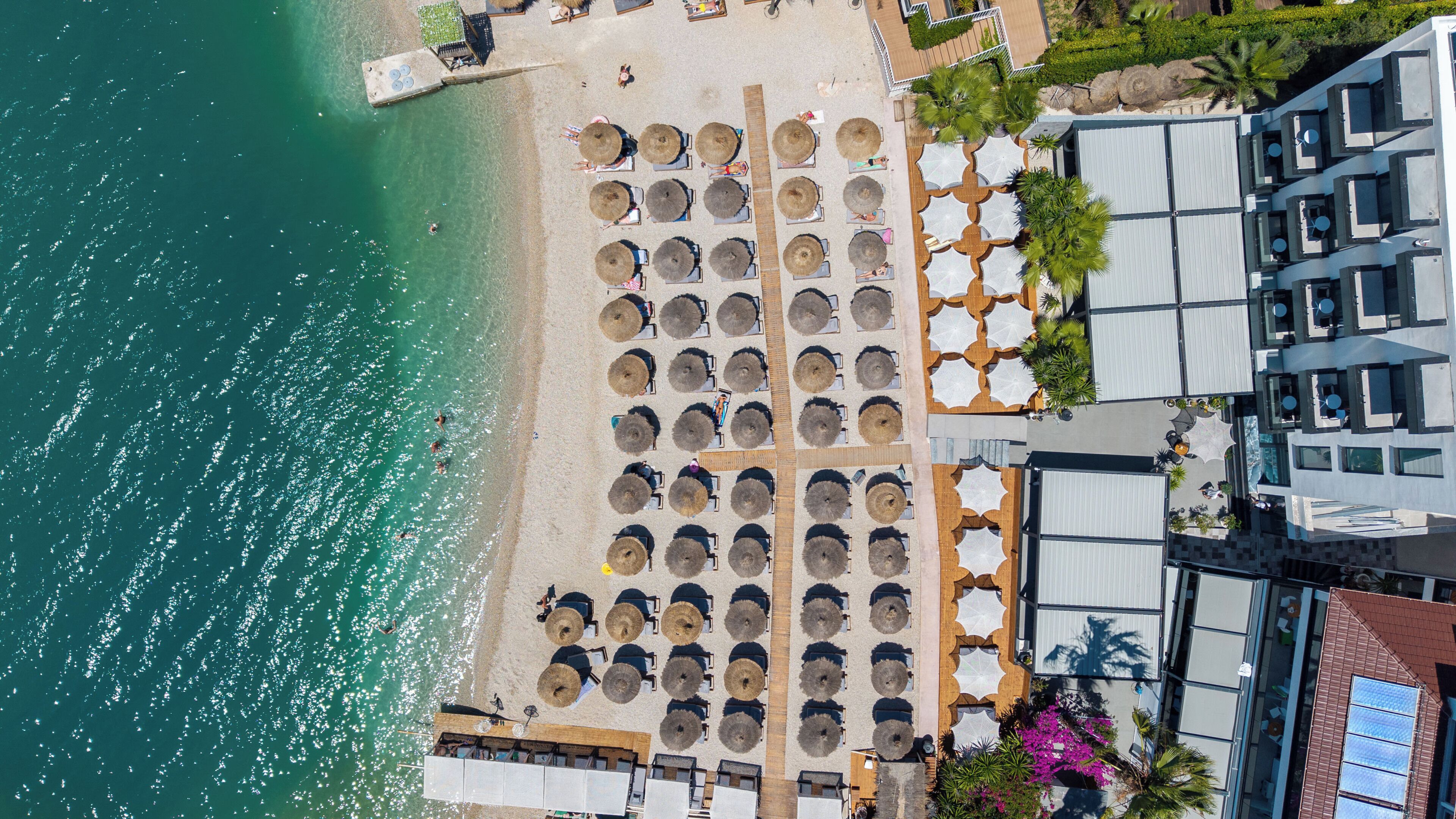 An aerial view of a beachside hotel with rows of sun loungers, a pool, and a building with multiple balconies.