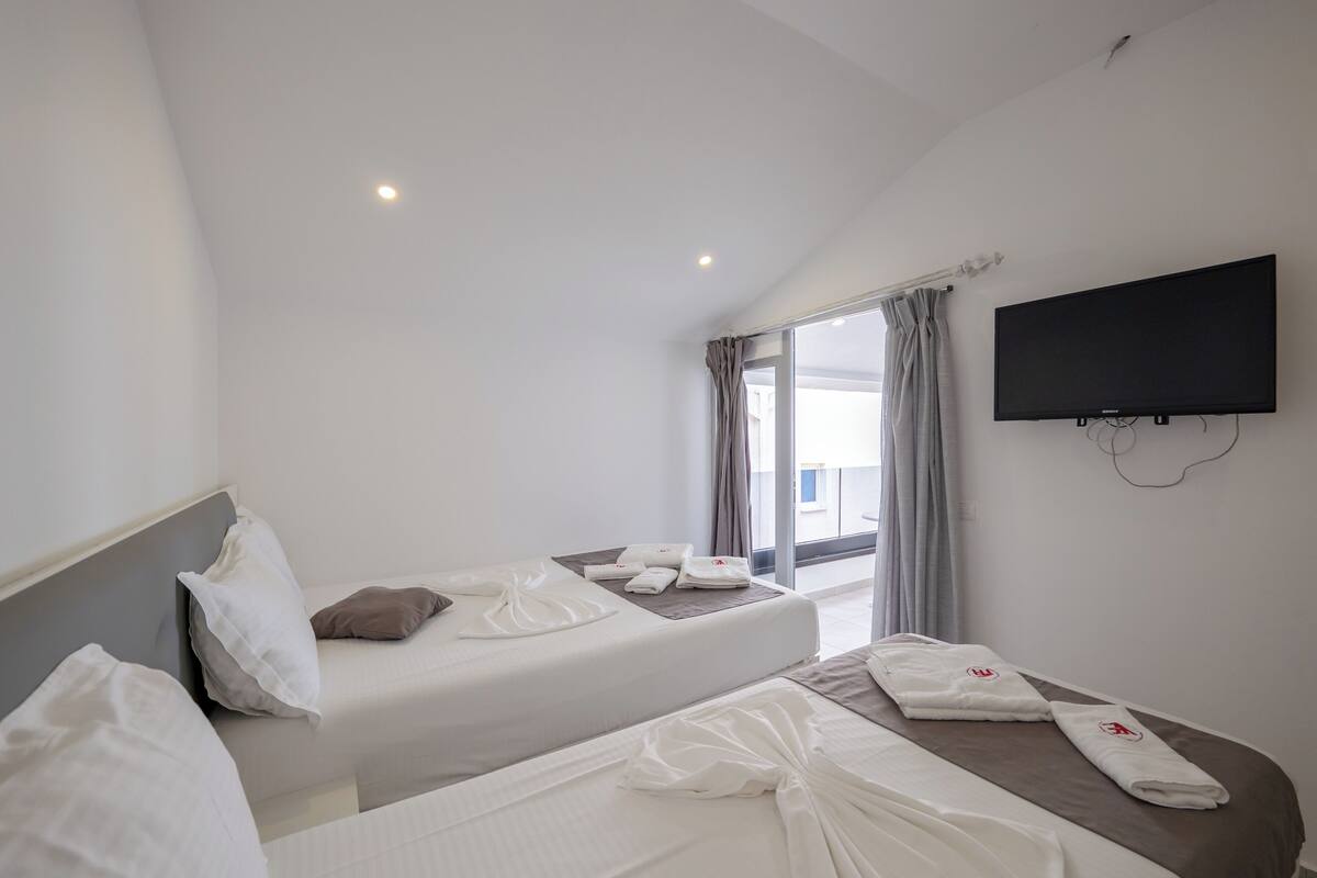 A hotel room with two beds, a flat-screen TV mounted on the wall, and a large window with curtains.