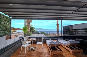 A modern outdoor lounge area with wooden flooring, white chairs, and sun loungers, overlooking a scenic ocean view.