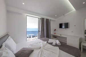 Una habitación de hotel moderna con una cama grande, un televisor de pantalla plana, un balcón con vistas al mar y un escritorio blanco con espejo.