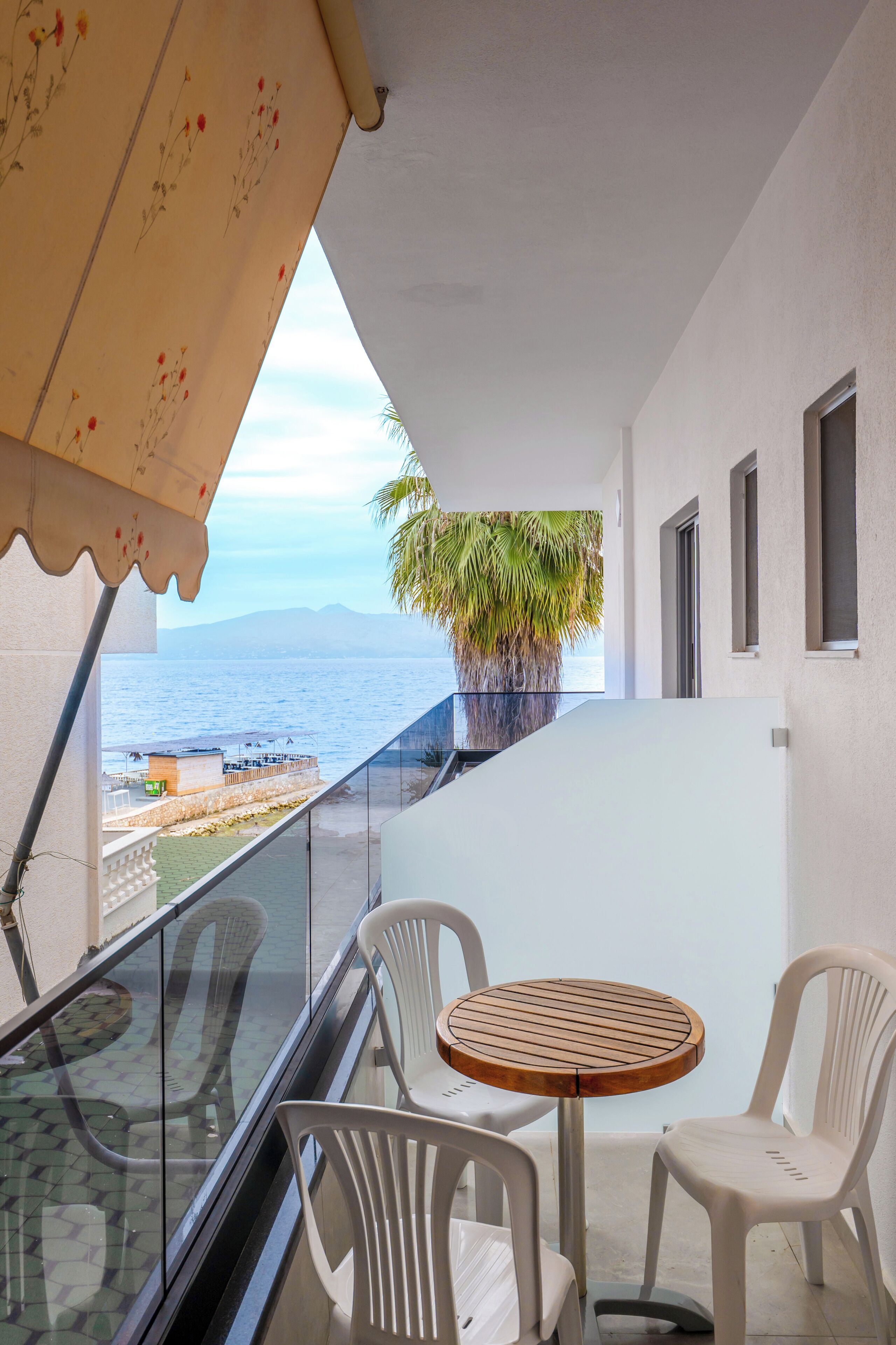 A balcony with a glass railing, a round wooden table, and white plastic chairs. A potted plant is visible in the background.