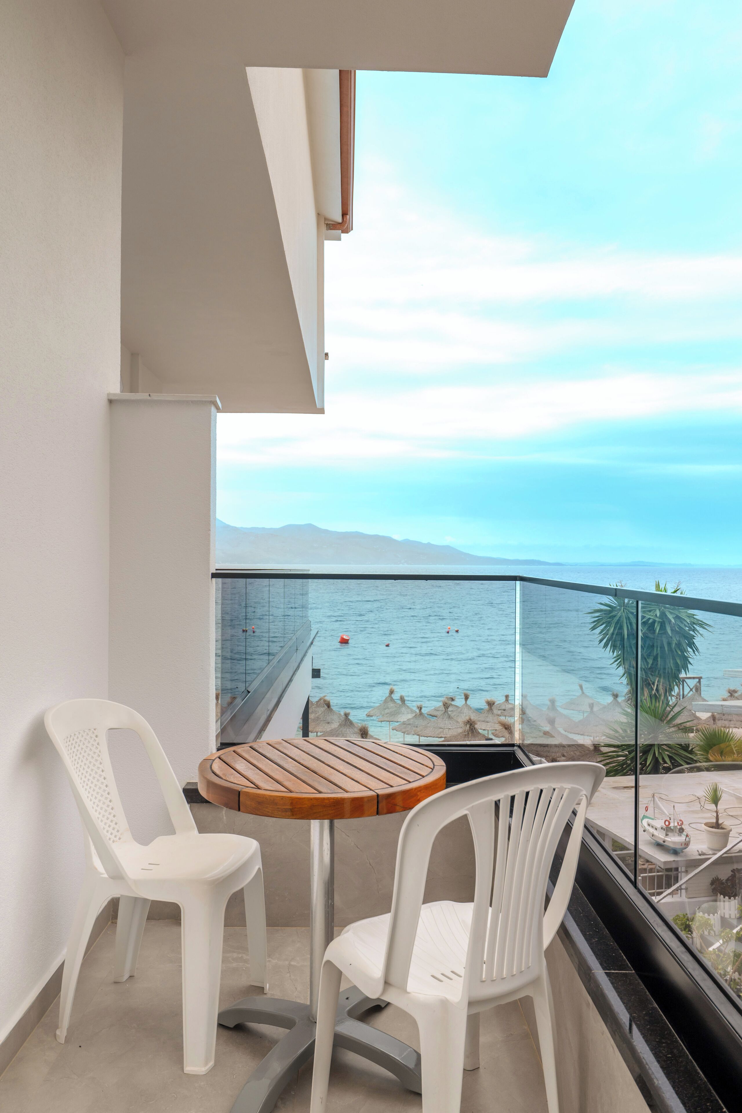 Un balcon avec deux chaises en plastique blanc et une table en bois, d’où l’on a une vue dégagée sur une plage, la mer et les montagnes lointaines.