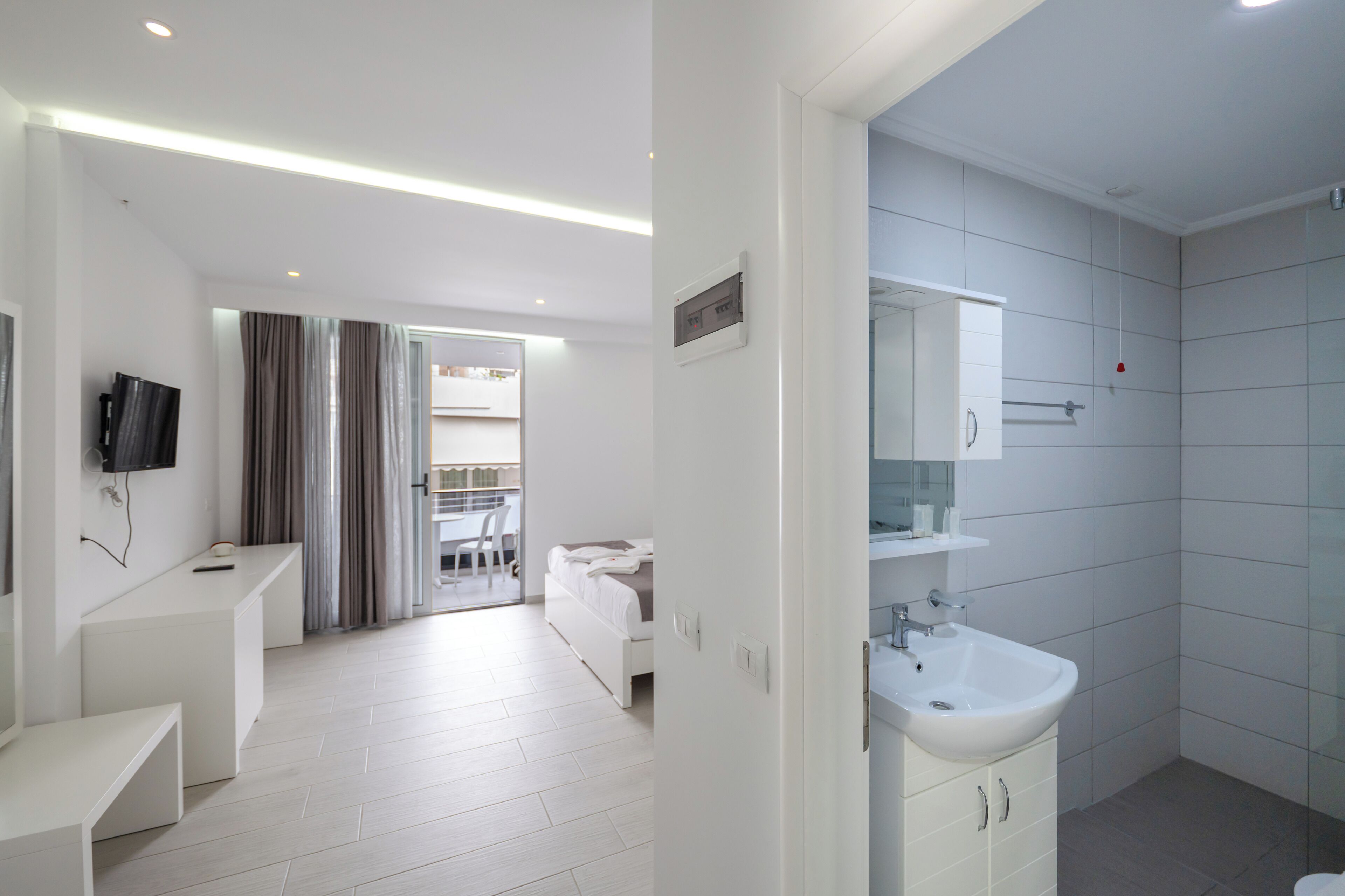 Ein modernes Hotelzimmer mit einem großen Bett, einem an der Wand befestigten Fernseher, einem Badezimmer mit Waschbecken und Spiegel sowie einem Balkon mit Vorhängen.