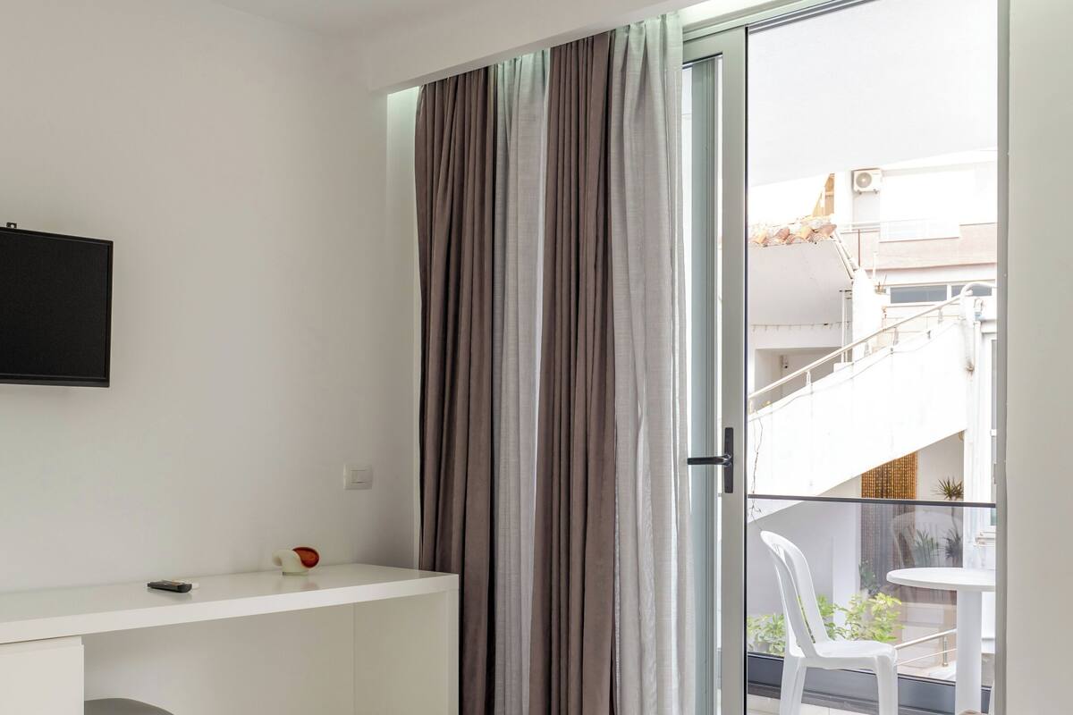 A modern hotel room with a bed, a desk, a TV, and a balcony with a view of the city.