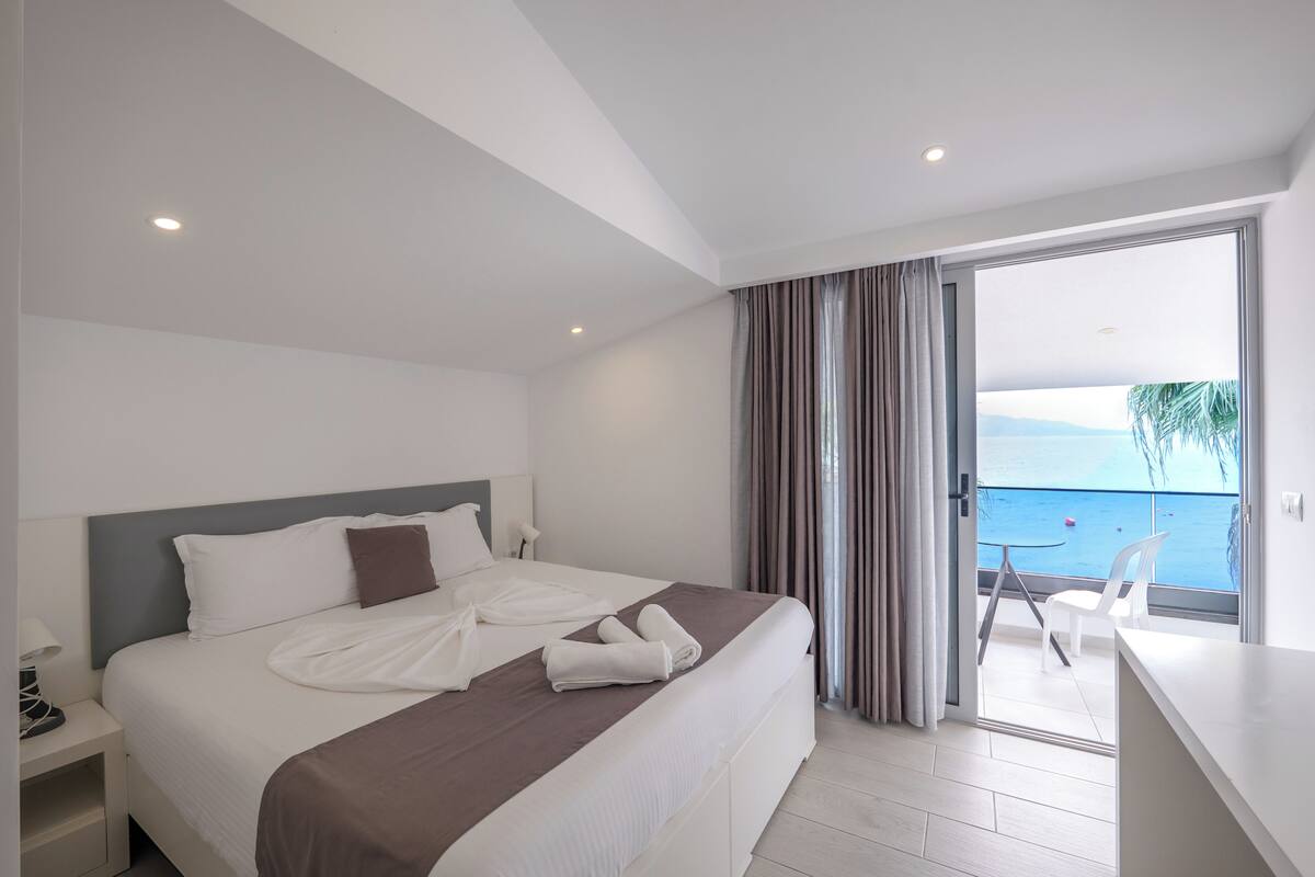 A modern hotel room with a large bed, a balcony with a view of the sea, and a small table with chairs.