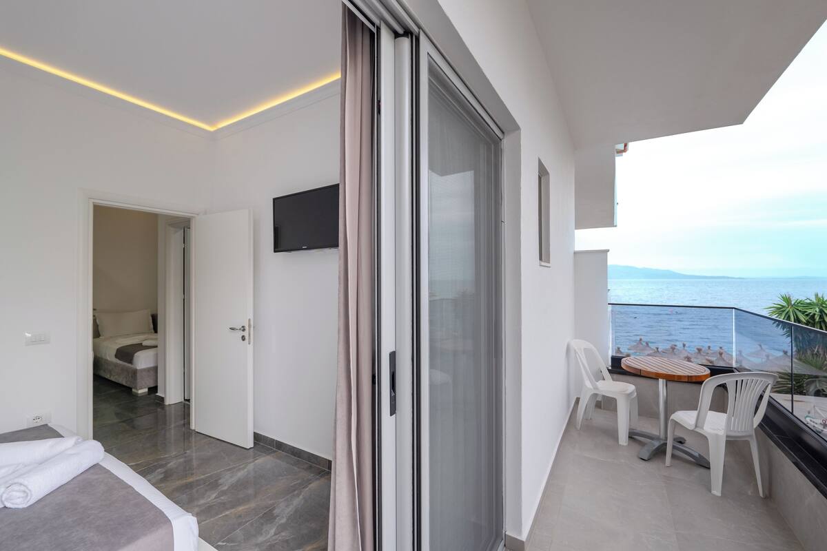 A modern balcony with a table and chairs, a sliding glass door, a flat-screen TV, and a view of the sea.