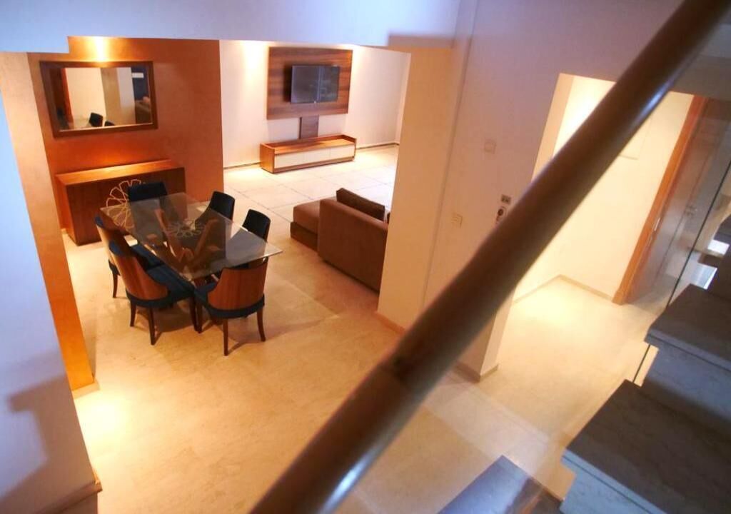 A modern living room with a glass dining table, a sofa, and a TV mounted on the wall.
