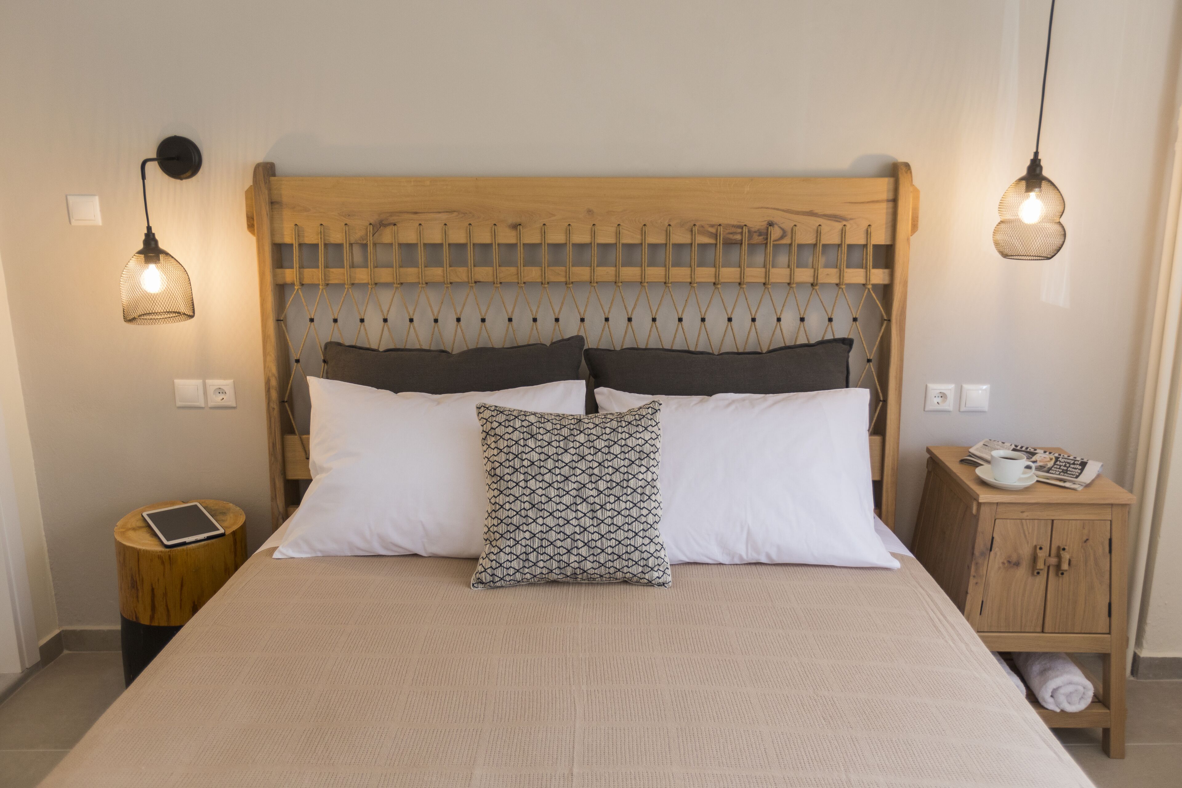 A neatly made bed with a patterned pillow, a wooden headboard, bedside tables with a tablet and a cup, and two hanging lamps.