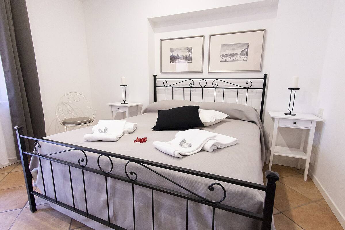 Un lit bien fait, avec du linge de lit blanc et une tête de lit noire, une table de chevet avec une lampe et une petite table de nuit blanche.
