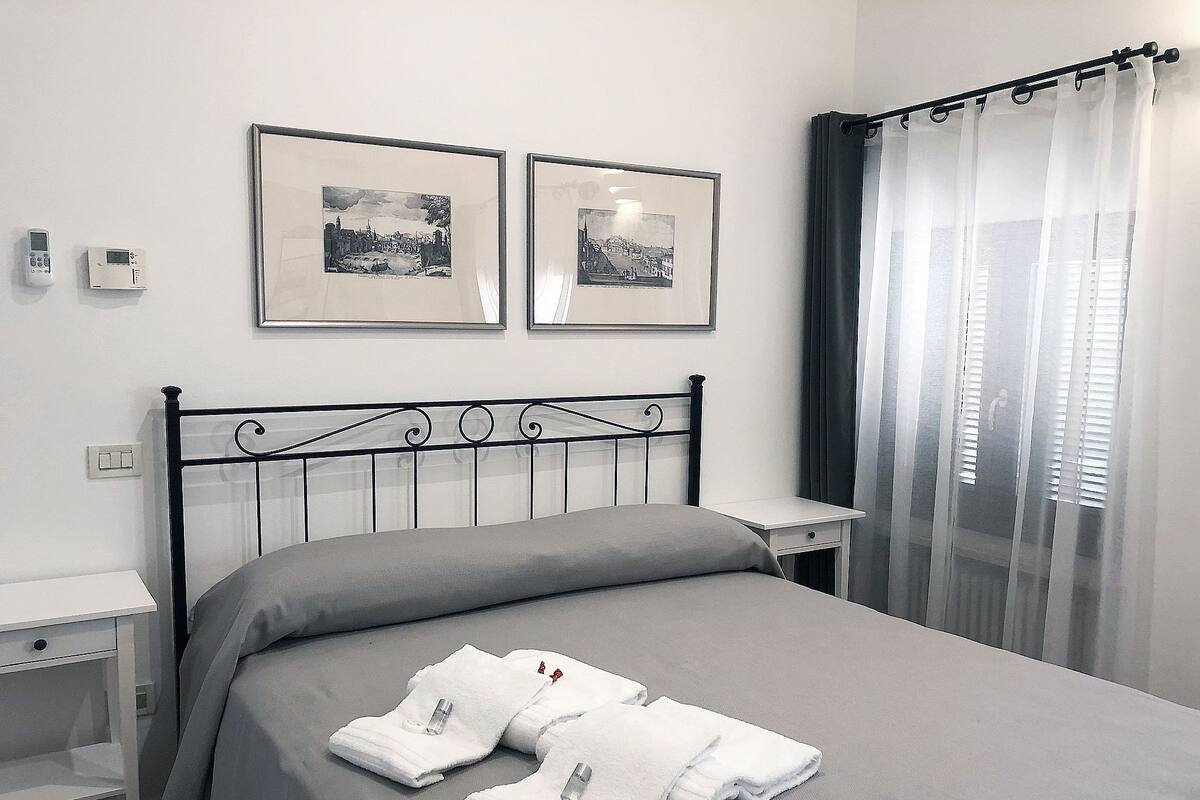 Un lit bien fait, recouvert d’une couverture grise, deux tableaux encadrés accrochés au mur, une table de chevet et une fenêtre avec un rideau.