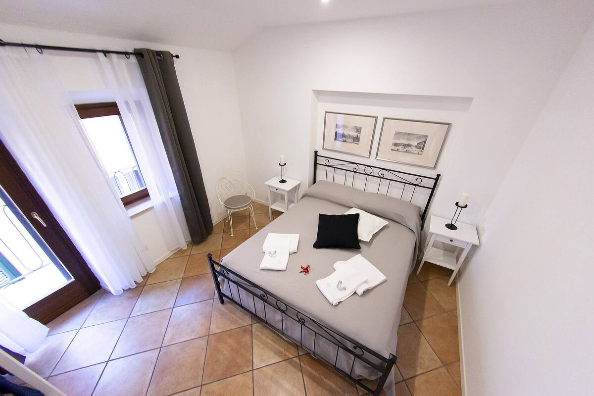 Un lit bien fait, avec du linge de lit blanc, un oreiller noir et un objet décoratif rouge. Il y a deux tables de chevet avec des lampes, une fenêtre avec des rideaux et deux tableaux encadrés accrochés au mur.