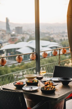 A dining table with various dishes and glasses of wine, set against a backdrop of a cityscape and pomegranate decorations. - Brim Hotel (Tbilisi)
