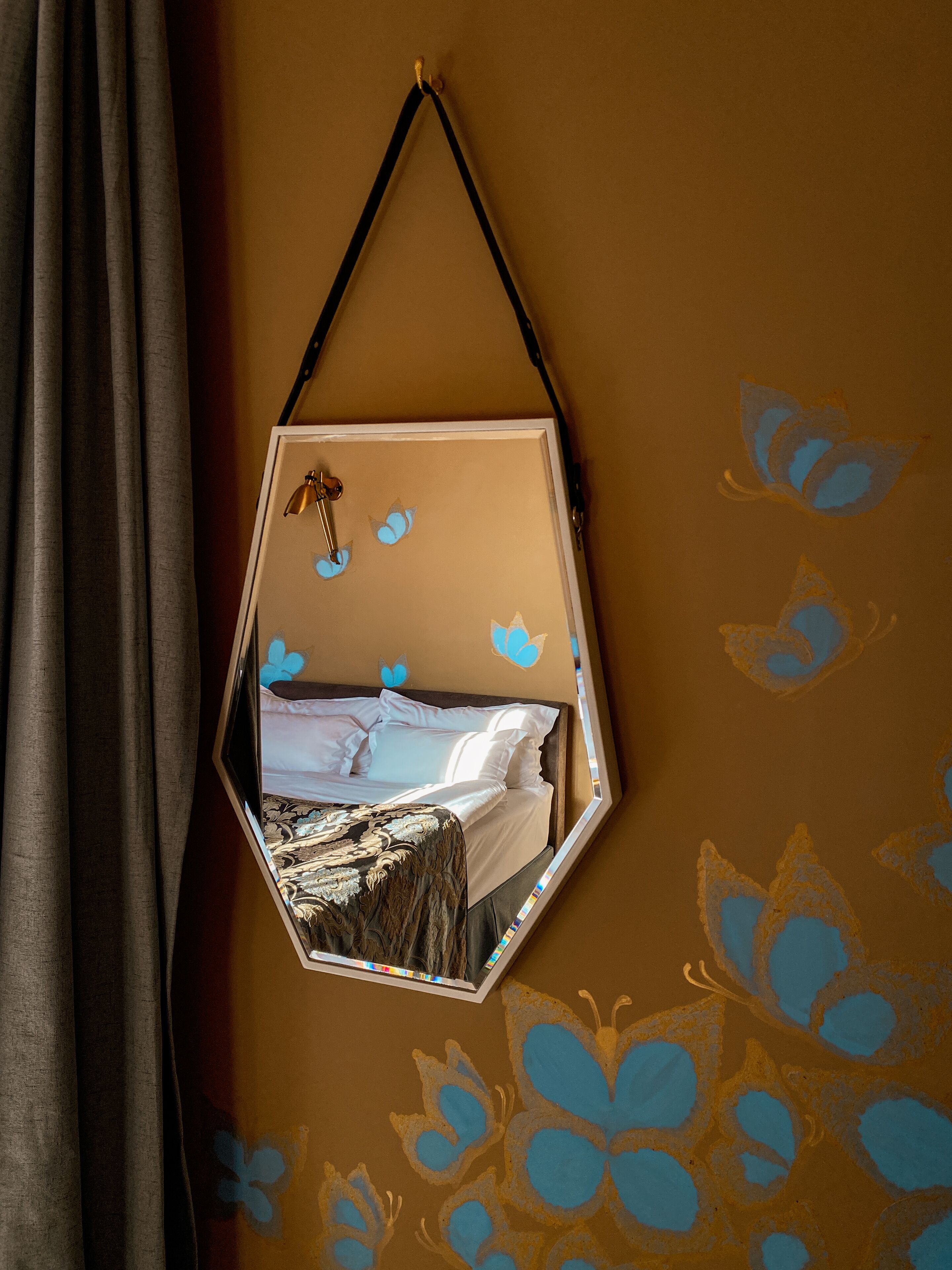 Een zeshoekige spiegel met weerspiegeling van een bed, vlinderdecoratie aan de muur en een gordijn.