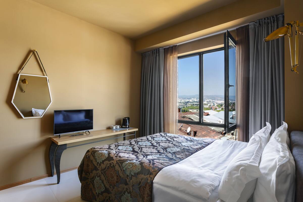 A hotel room with a bed, a TV, a desk, and a large window with a city view.