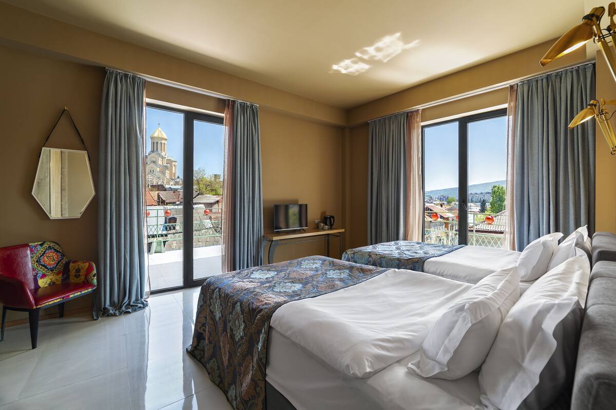 A hotel room with two beds, a red bench, a TV, and a view of a cityscape.