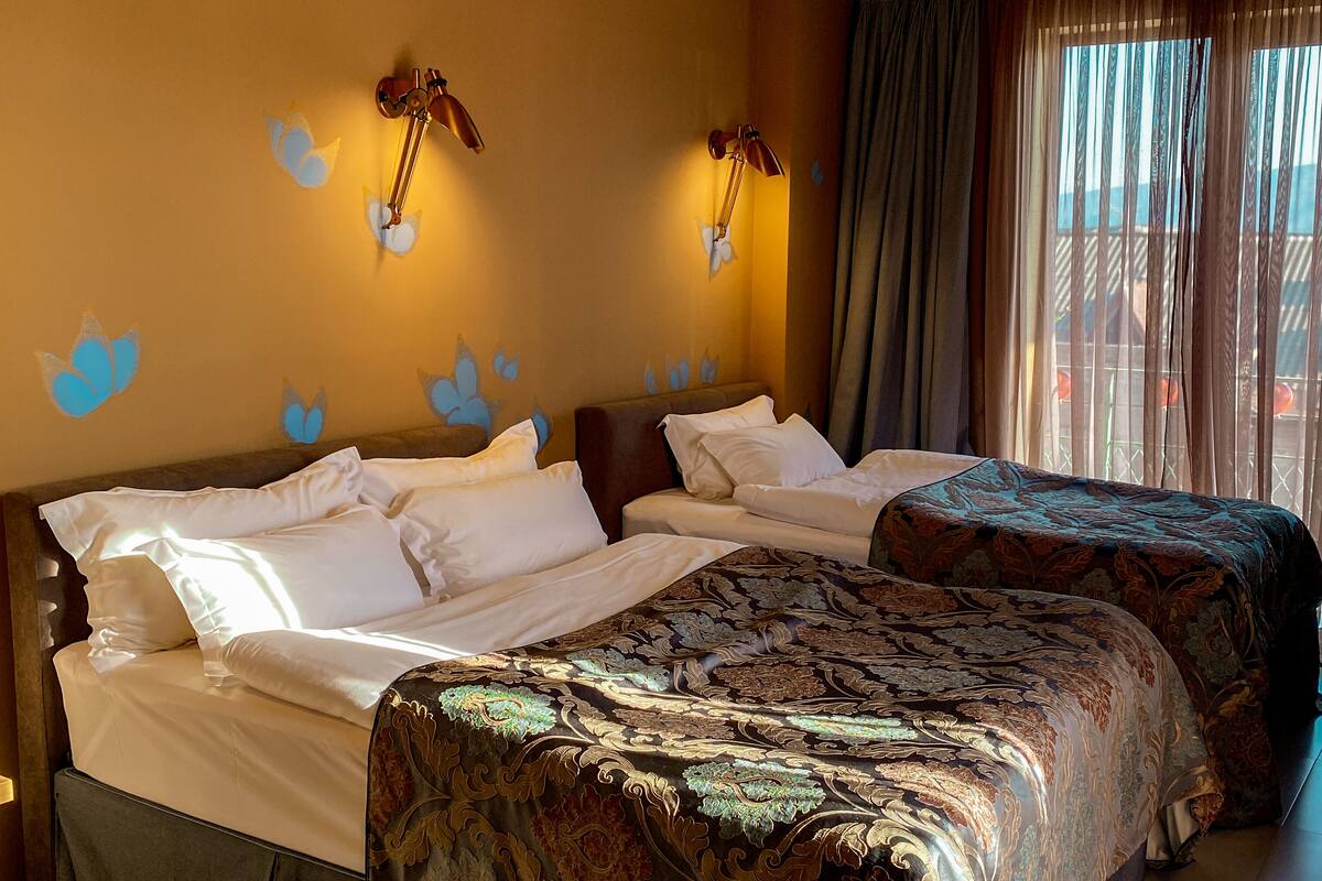 A hotel room with two beds, a large window with curtains, and wall-mounted lamps.