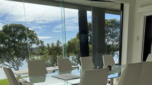 Un comedor moderno con una mesa de vidrio y sillas blancas, ventanas amplias con vistas a árboles y un lago, y suelo de baldosas.