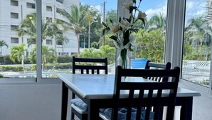 Un comedor con mesa y sillas, ventanas amplias con vista a palmeras y edificios, y un toldo azul.