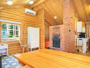 L’intérieur d’une cabane en rondins, avec une table en bois, une armoire blanche, une cheminée en briques et une fenêtre donnant sur un paysage verdoyant.